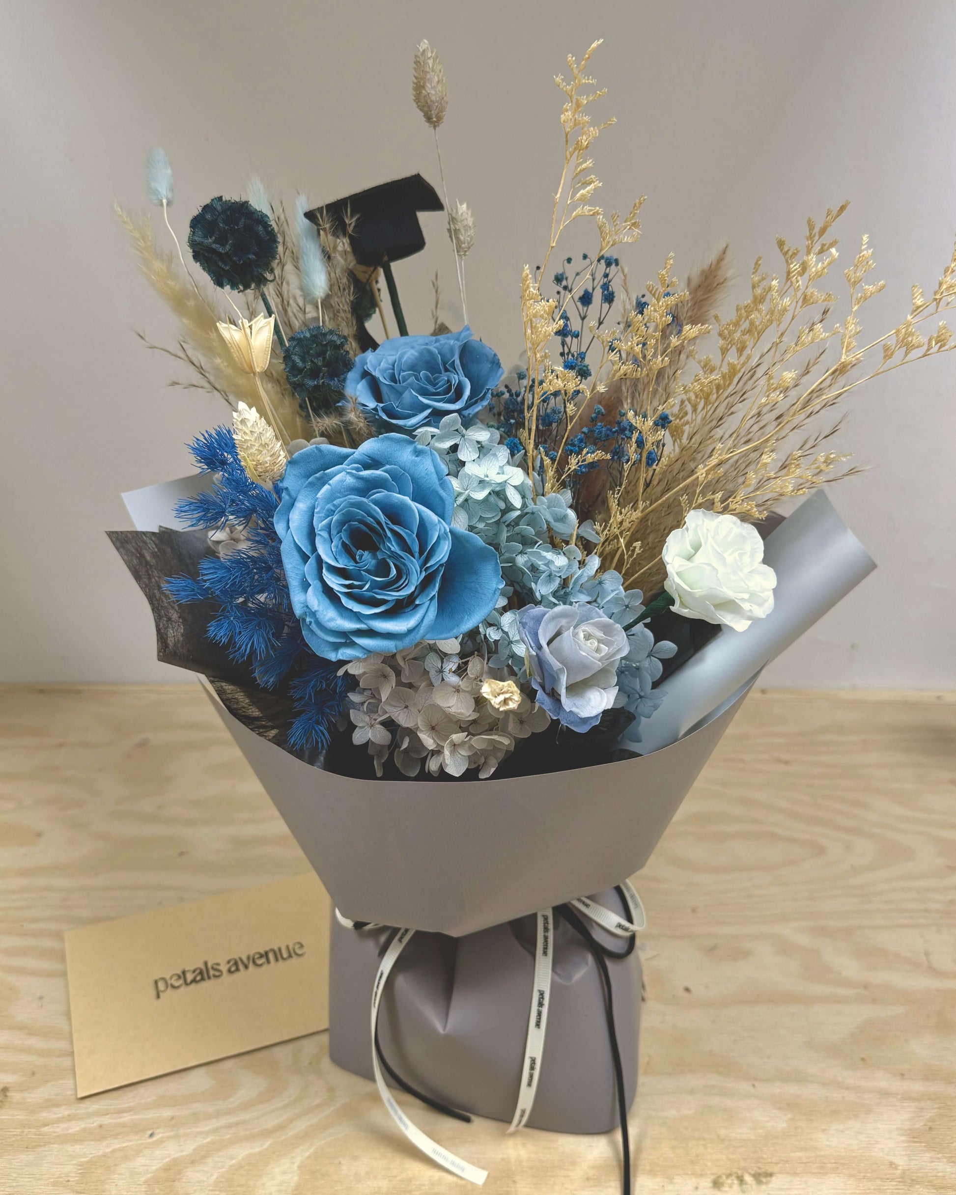 Bouquet of blue and white preserved  flowers in a gray wrap with a card on a wooden surface.